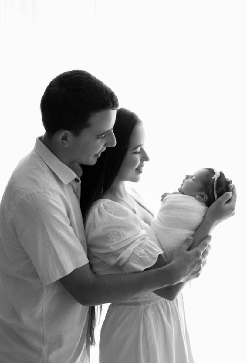 Black and white photo of parents lovingly gazing at their newborn baby wrapped in white cloth.