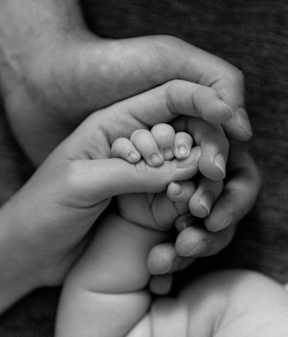 Close-up black and white photo of adult hands gently holding a newborn baby's tiny hand.