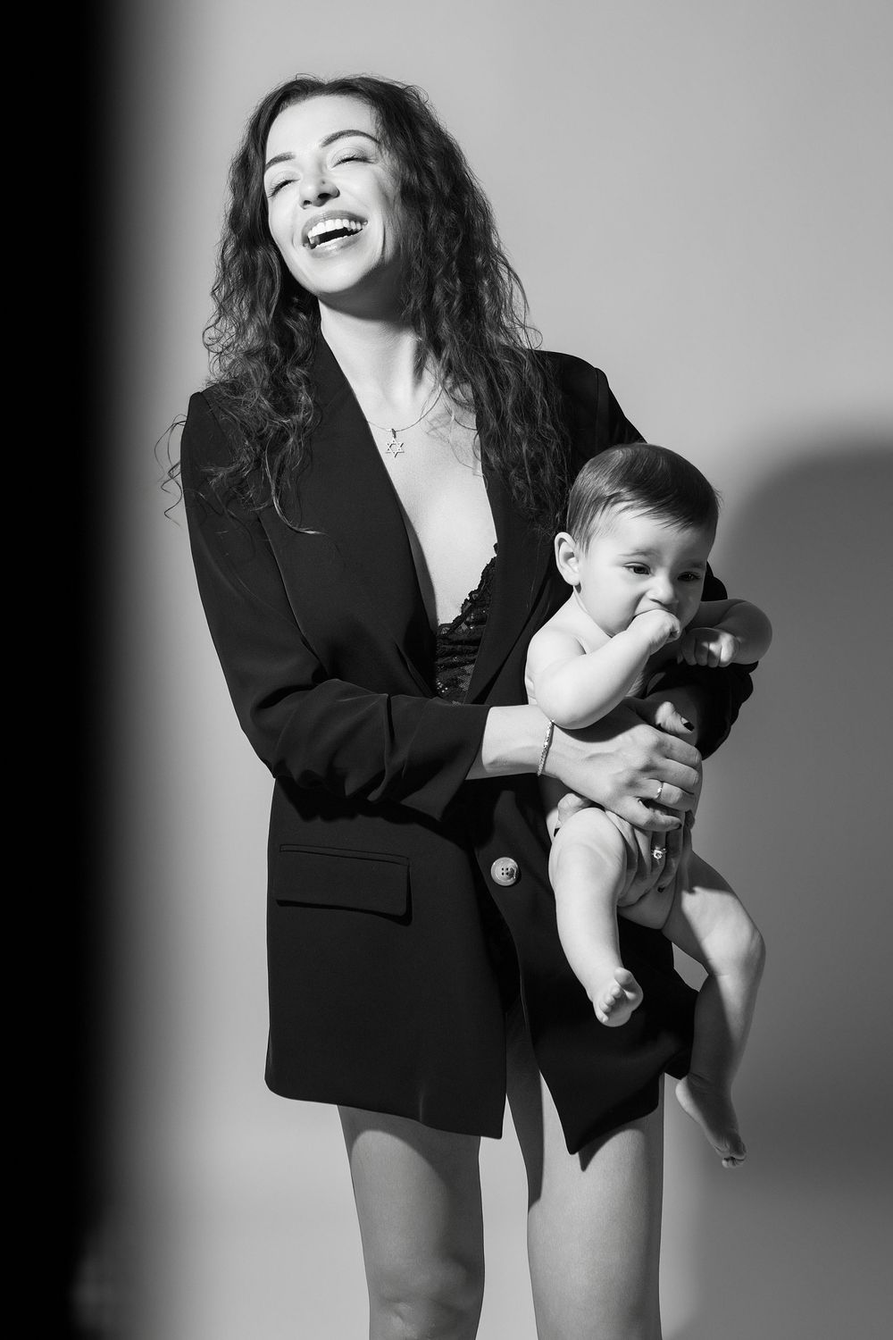 Black and white photo of a laughing woman in a blazer holding a cute baby against a light background.