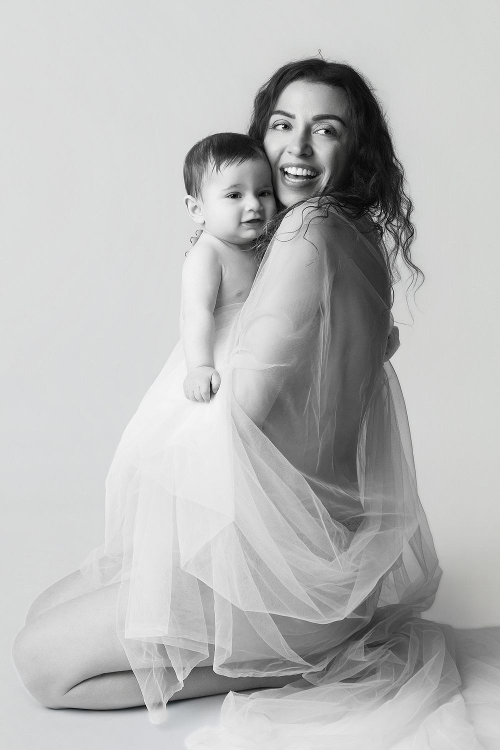 Black and white photo of a smiling mother in sheer white dress holding her baby while kneeling on a studio floor.