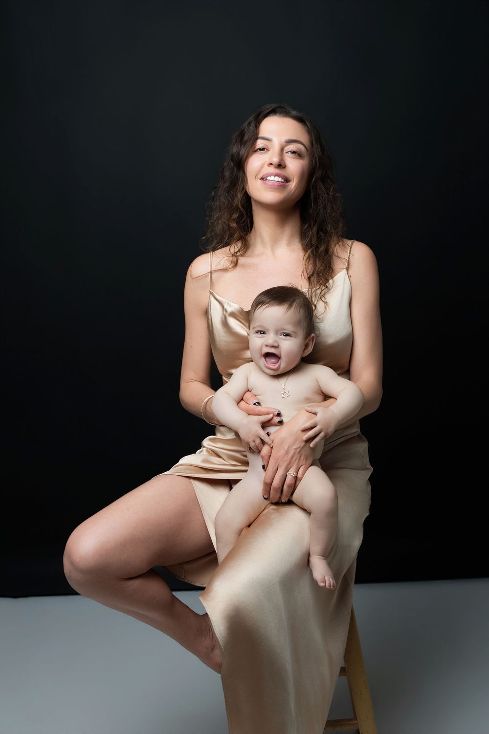 A smiling young mother in a gold satin dress sits holding her laughing baby against a dark background.