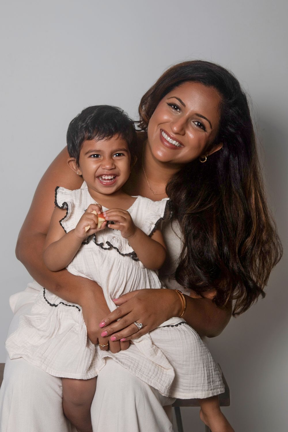 Smiling woman holding a happy toddler, both wearing white outfits, posing together for a portrait photo.