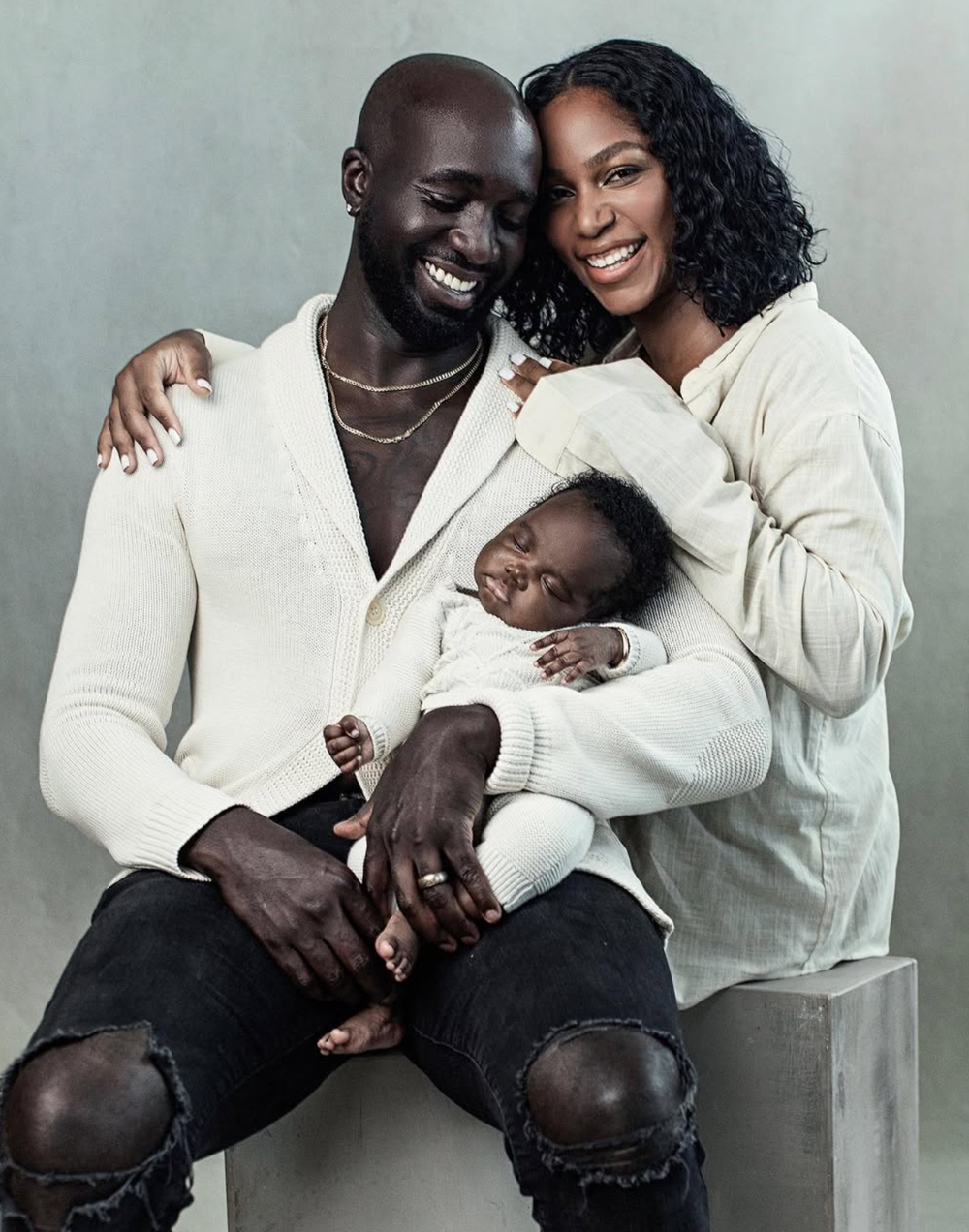 A happy family portrait with a couple in white outfits holding their sleeping newborn baby.