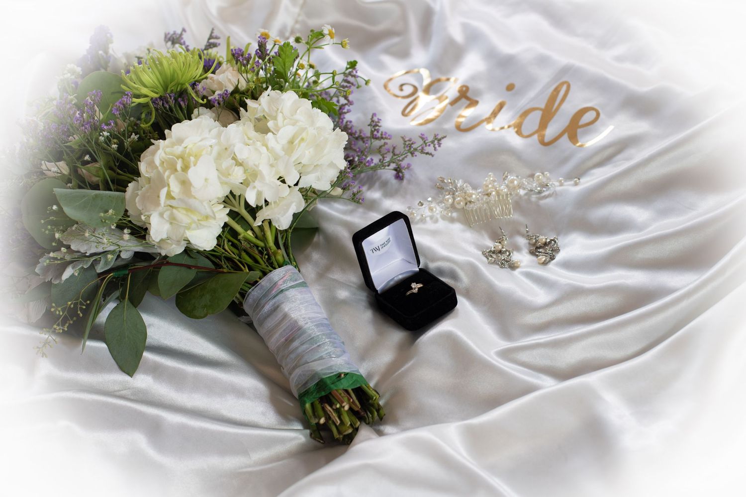 Wedding bouquet with white hydrangeas and greenery next to engagement ring box on white satin fabric.