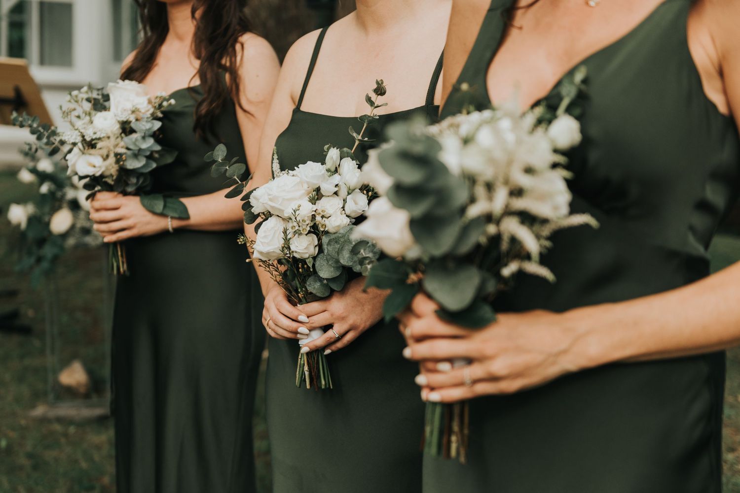Bridesmaids in elegant green dresses hold white and green floral bouquets during outdoor wedding ceremony.
