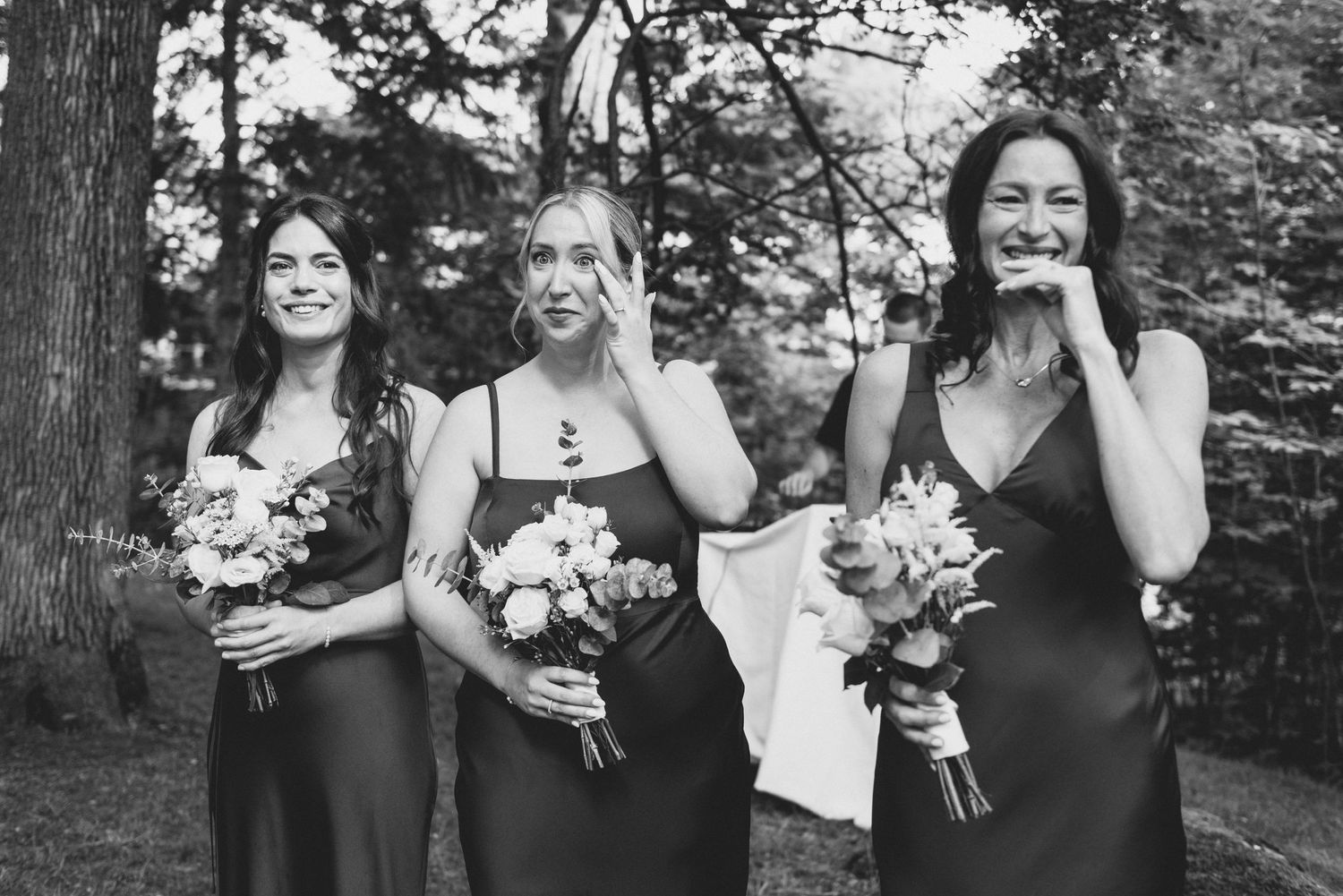 Three bridesmaids in black dresses hold bouquets while sharing joyful laughter in a forest setting.
