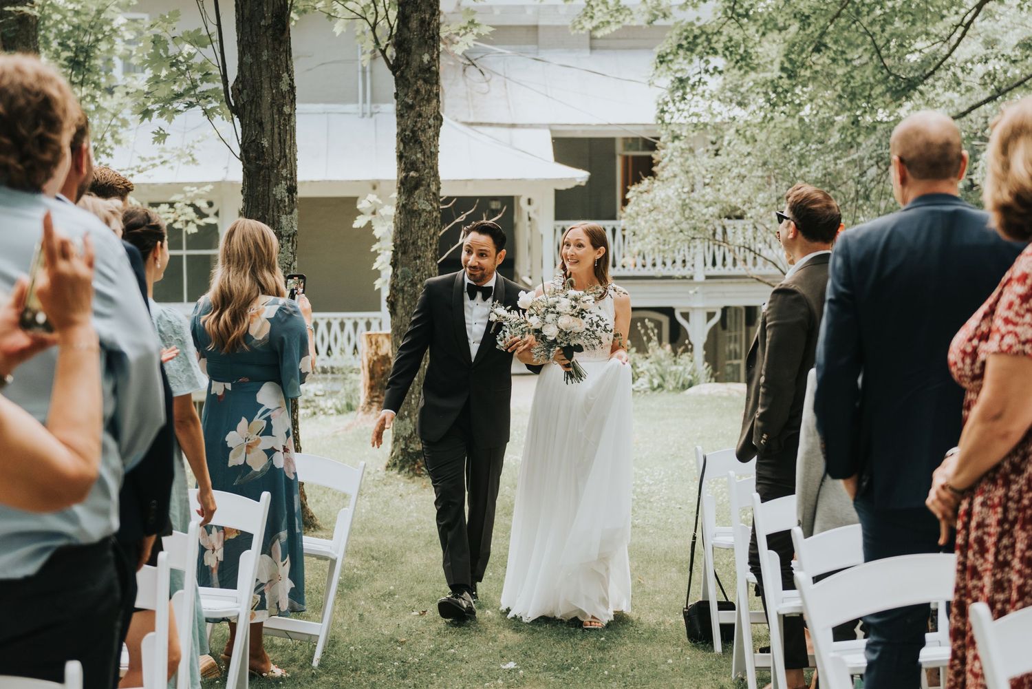 A couple walks down the aisle together at an outdoor garden wedding ceremony with guests seated on white chairs.