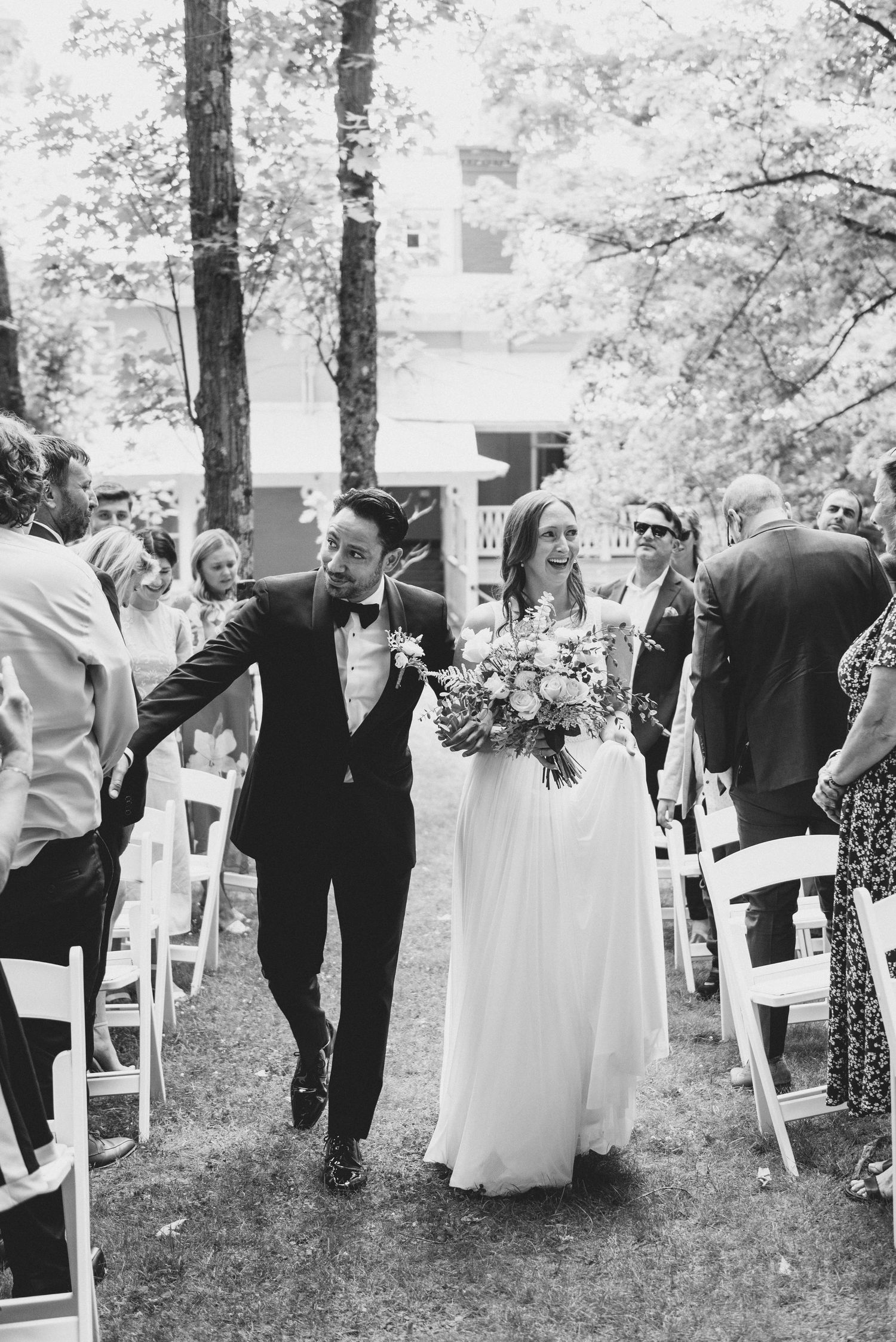 A black and white wedding ceremony photo showing a couple walking down the outdoor aisle in a wooded setting.
