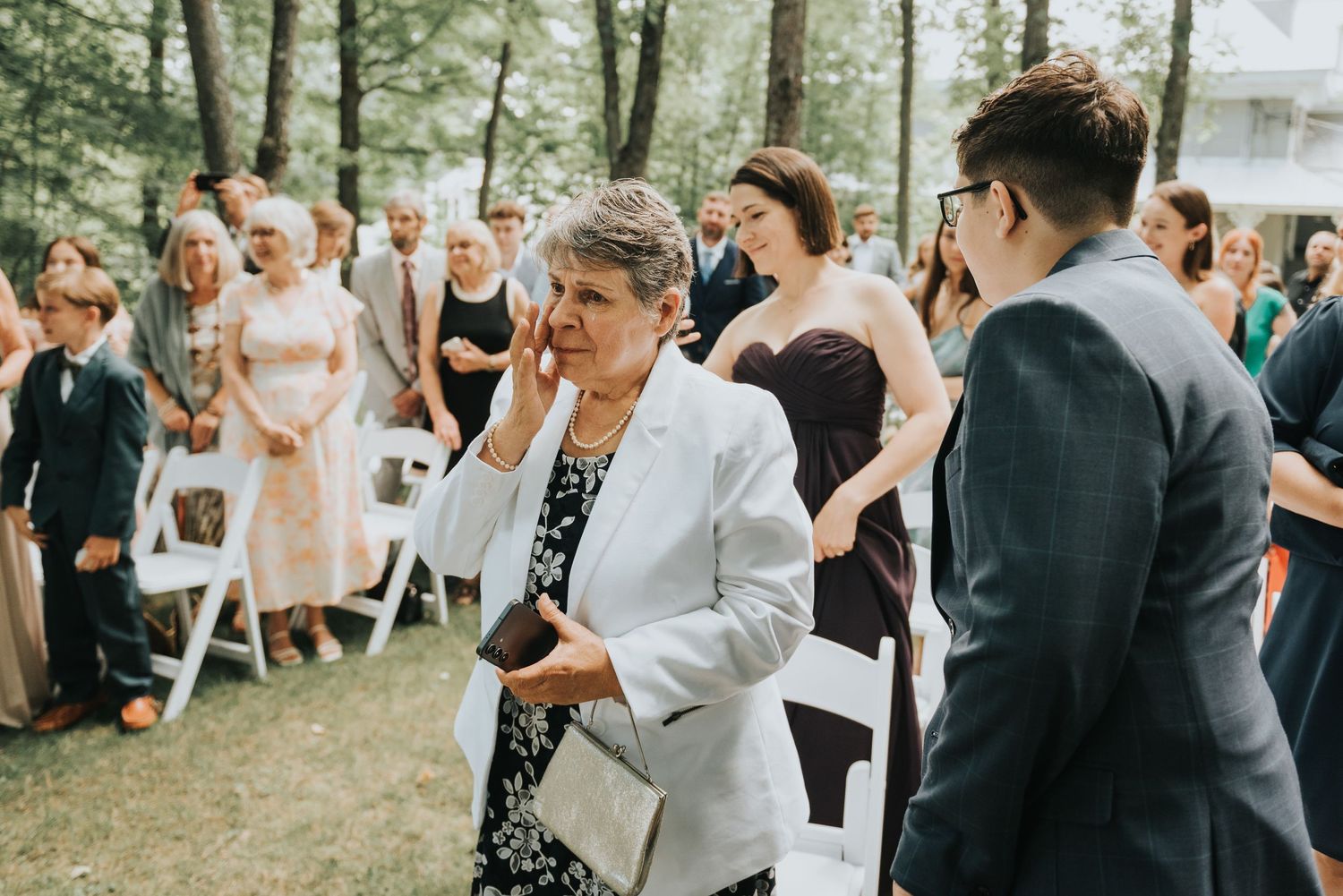 Guests stand together at an outdoor wedding ceremony beneath trees on a bright summer day.