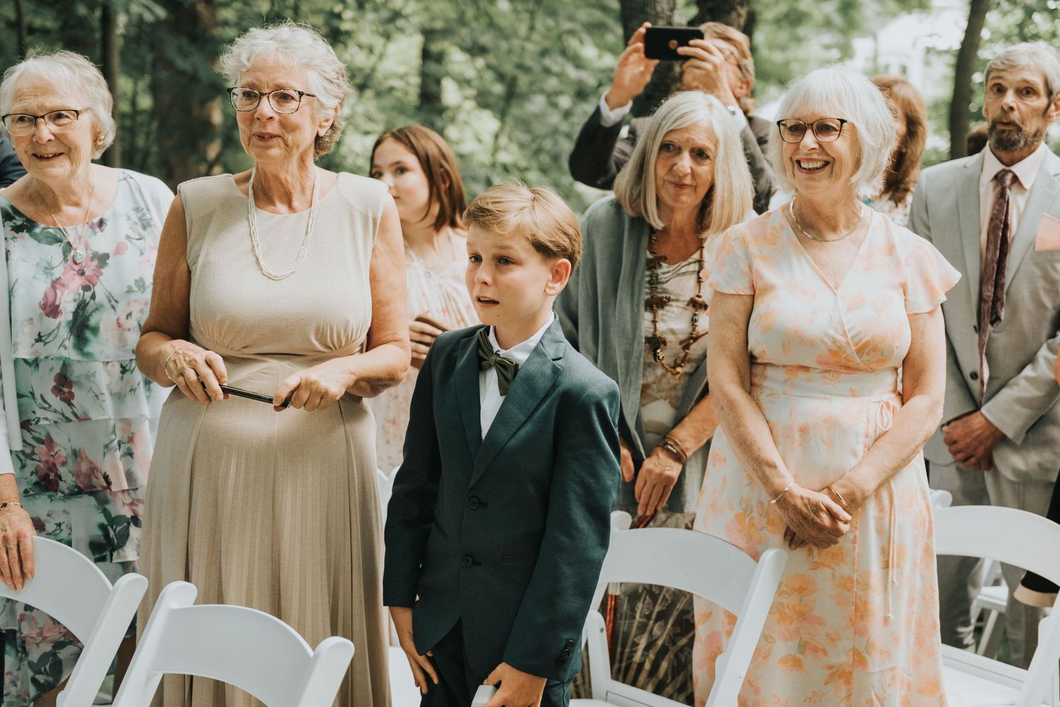 Wedding guests and family members stand together during an outdoor ceremony among trees.