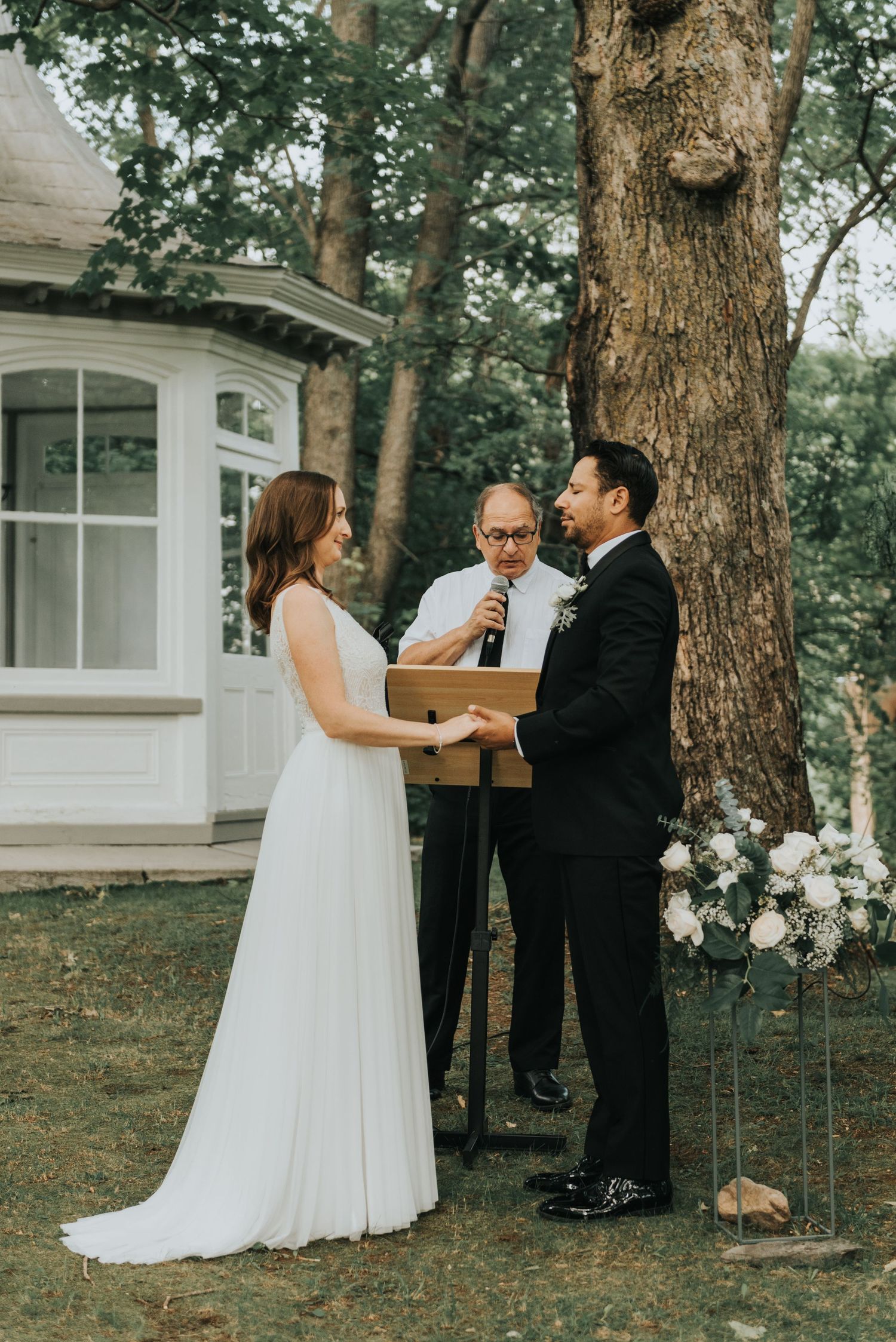 A couple exchanges vows during their outdoor wedding ceremony in front of a white gazebo and large tree.