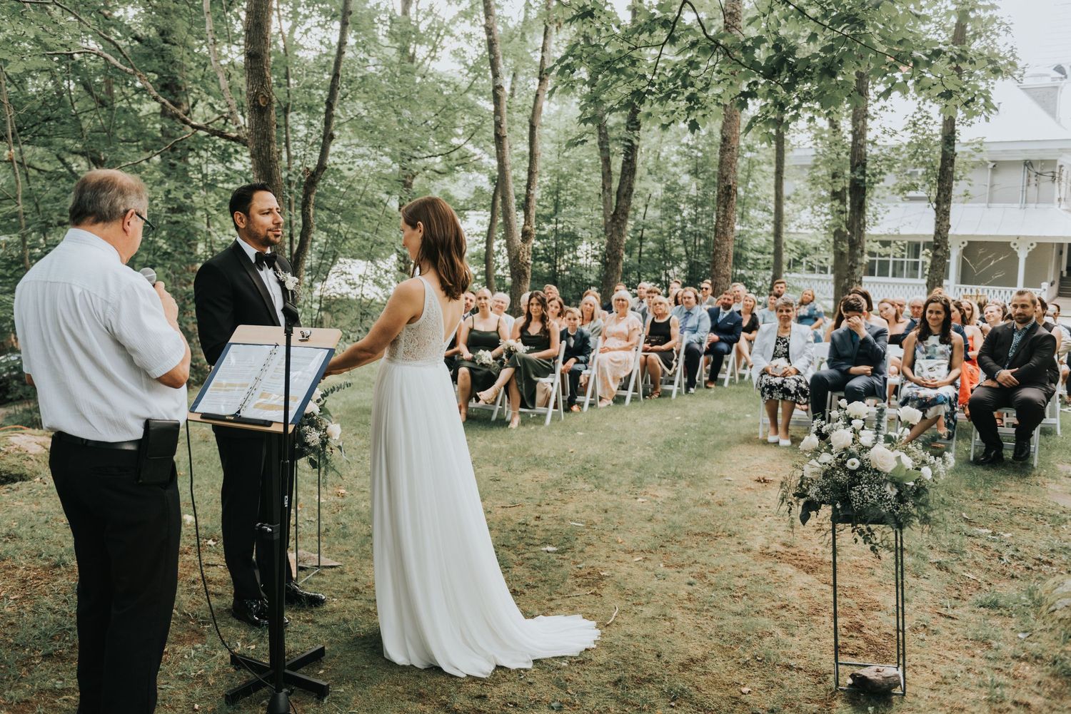 An outdoor wedding ceremony in a wooded setting with guests seated on white chairs watching the couple exchange vows.