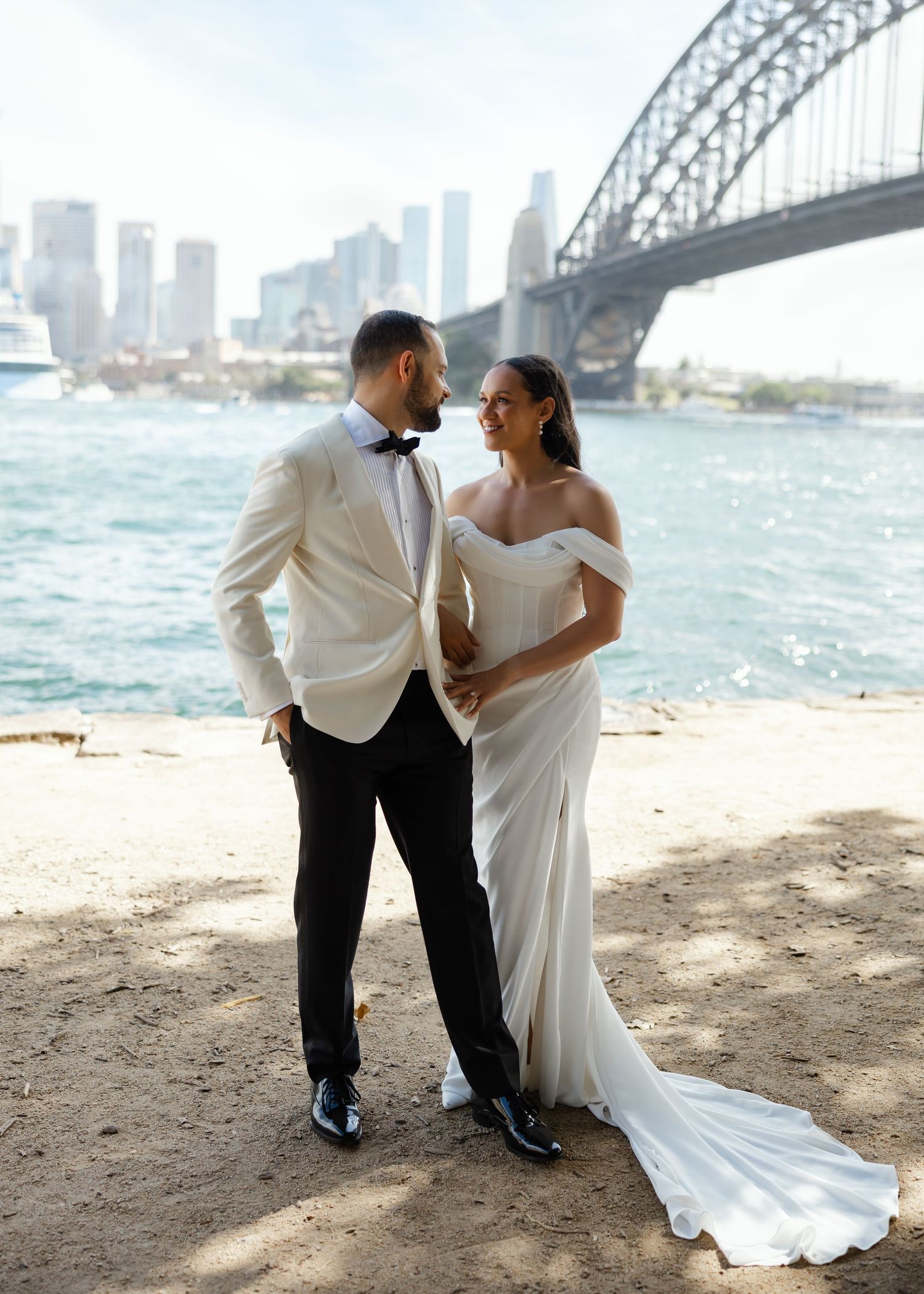Bride and groom pose near Sydney Harbour Bridge waterfront, she in off-shoulder gown, he in white tuxedo jacket.