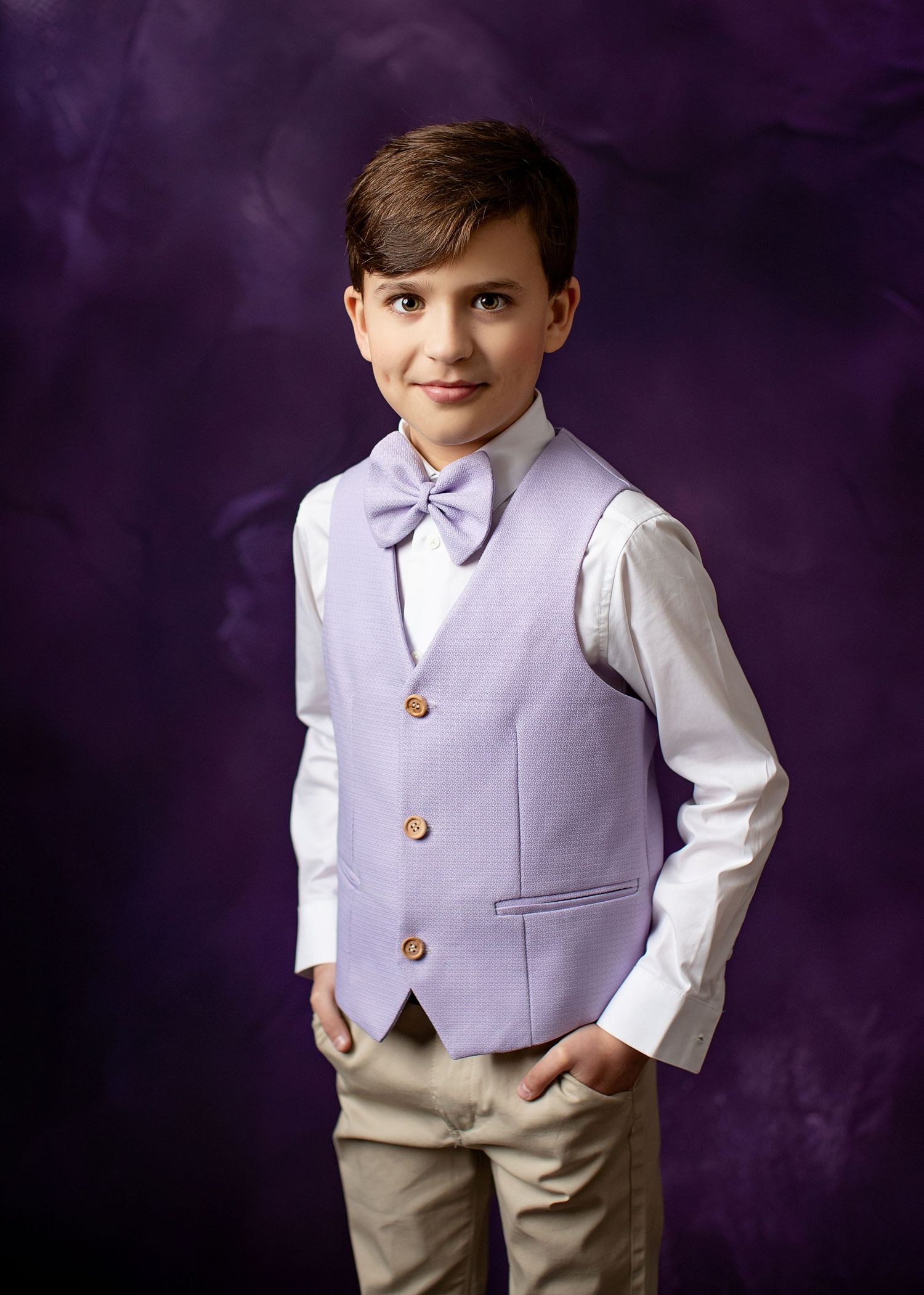 Young boy wearing a lavender vest, white shirt, purple bowtie and khaki pants poses against a purple backdrop.