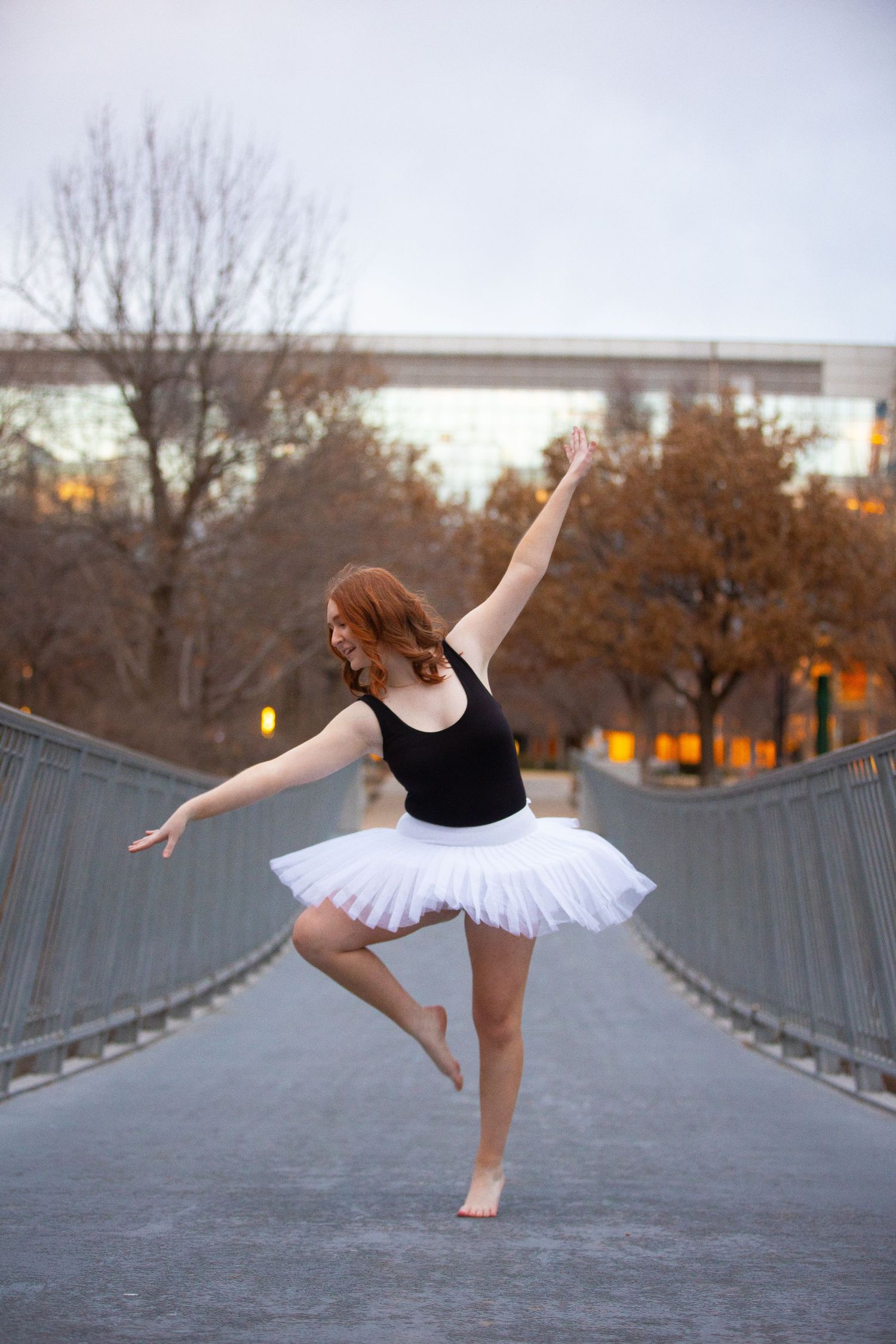 Ballet dancer in white tutu performs graceful pose on bridge at dusk with autumn trees in background.