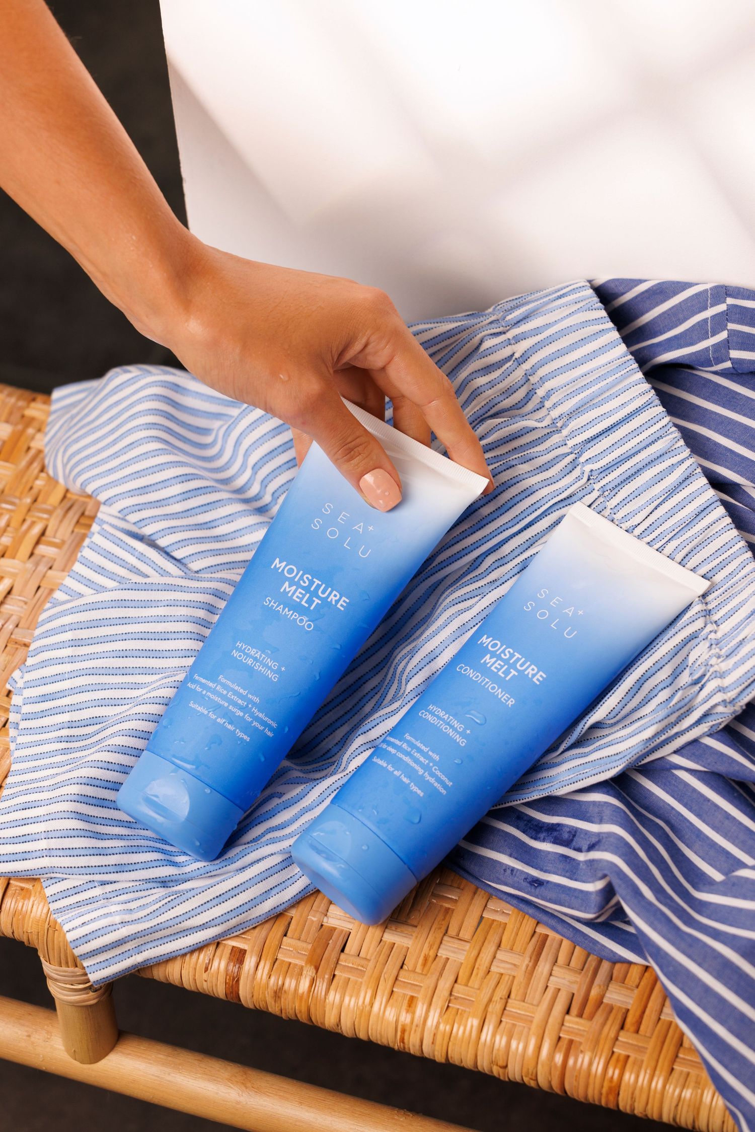 Two blue Shampoo & Conditioner tubes displayed on striped towel and woven surface. A wet hand is reaching for them.