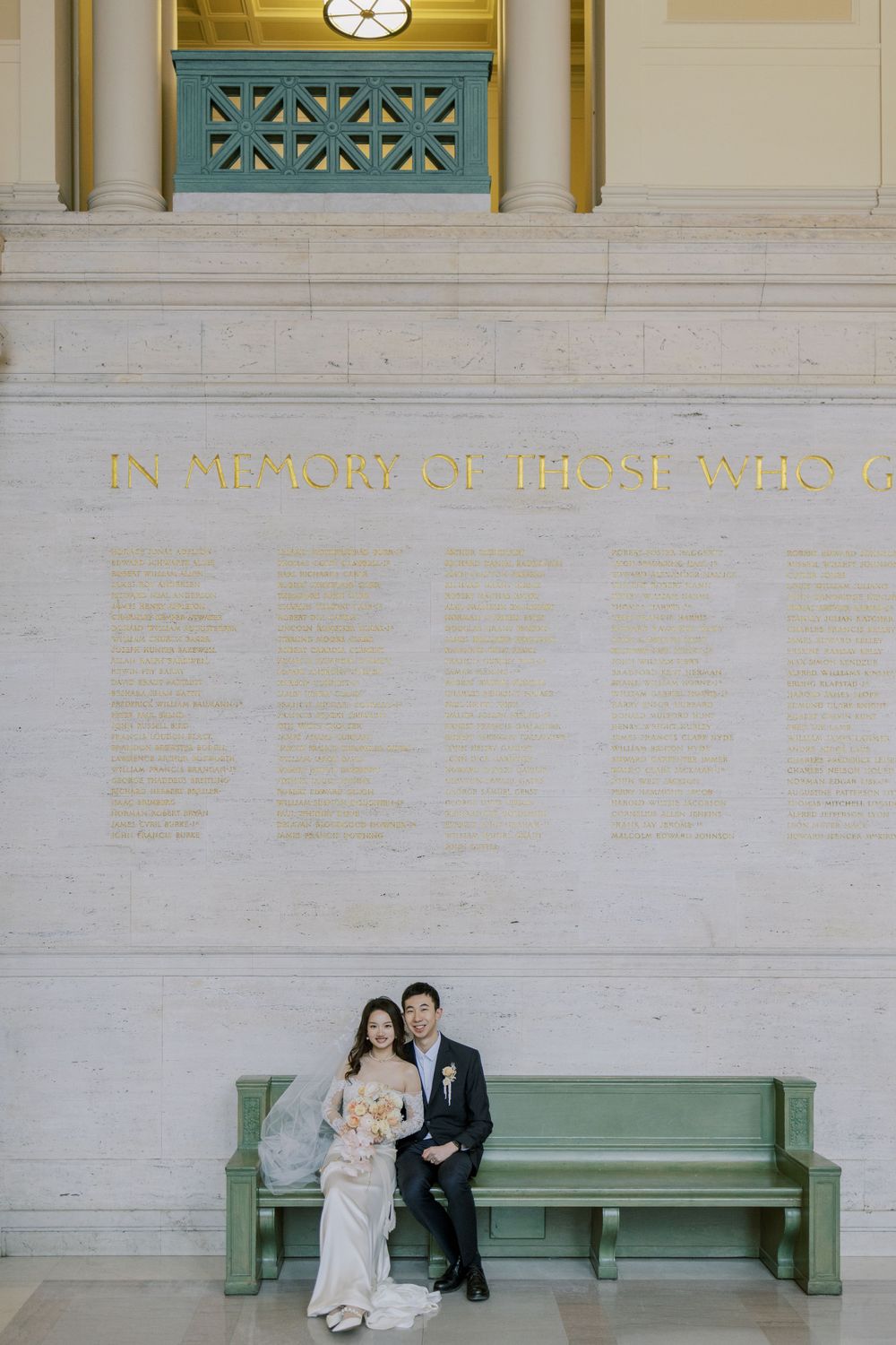 A couple sits on a green bench beneath a memorial wall inscription.