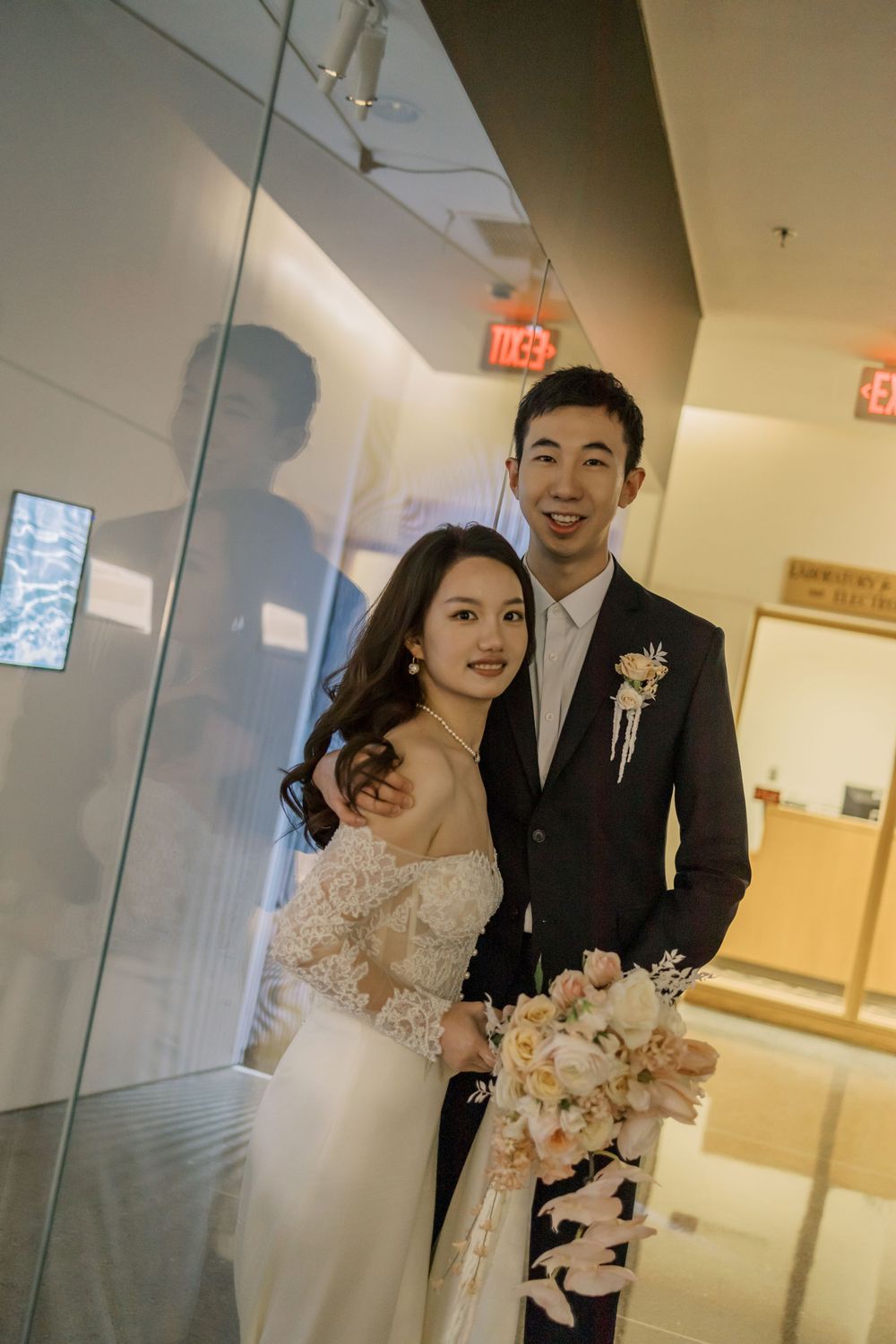 A wedding couple poses in a modern hallway with glass walls and bright lighting.