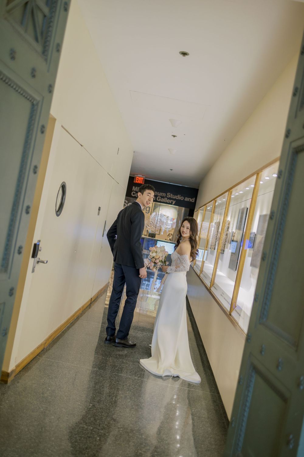 A wedding couple stands in a long institutional hallway with display cases along the wall.