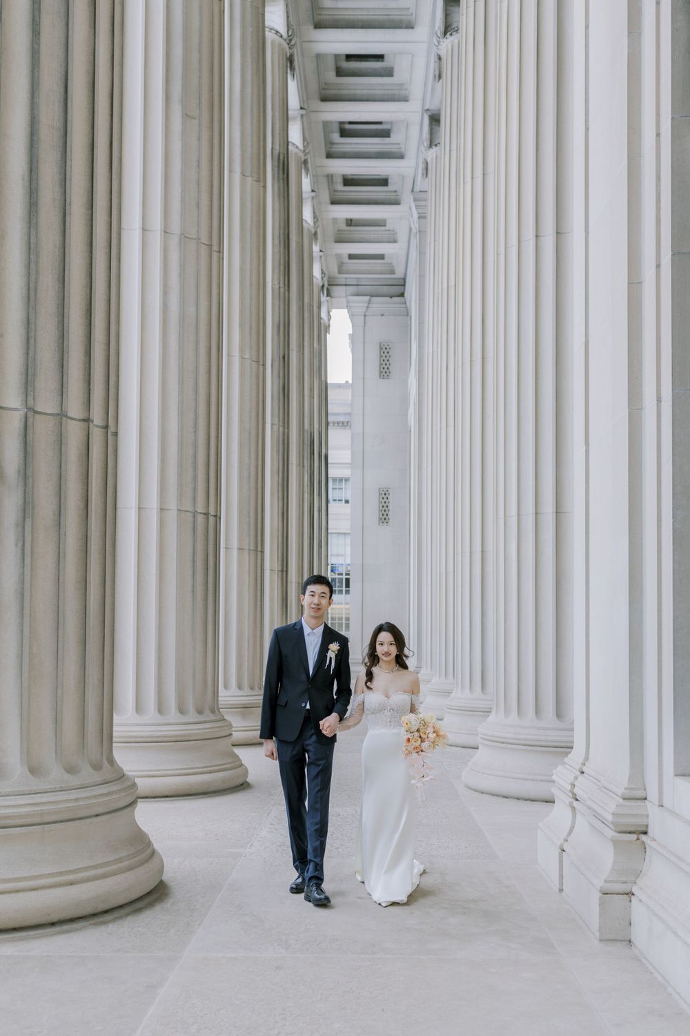Wedding couple walks amidst grand white marble columns of a classical building.