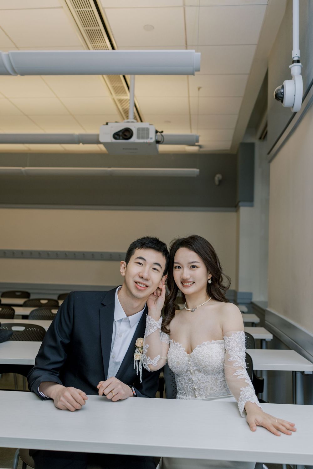 Wedding couple sits at classroom desk under ceiling-mounted projector.