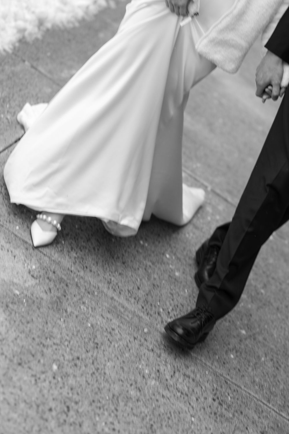 Black and white photo showing formal shoes and dress hem on concrete ground.
