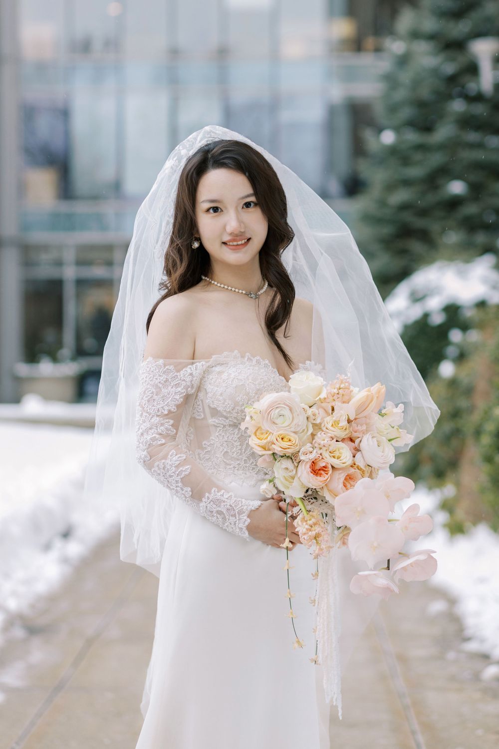 Winter bride in lace off-shoulder dress holds peach bouquet against snowy backdrop.