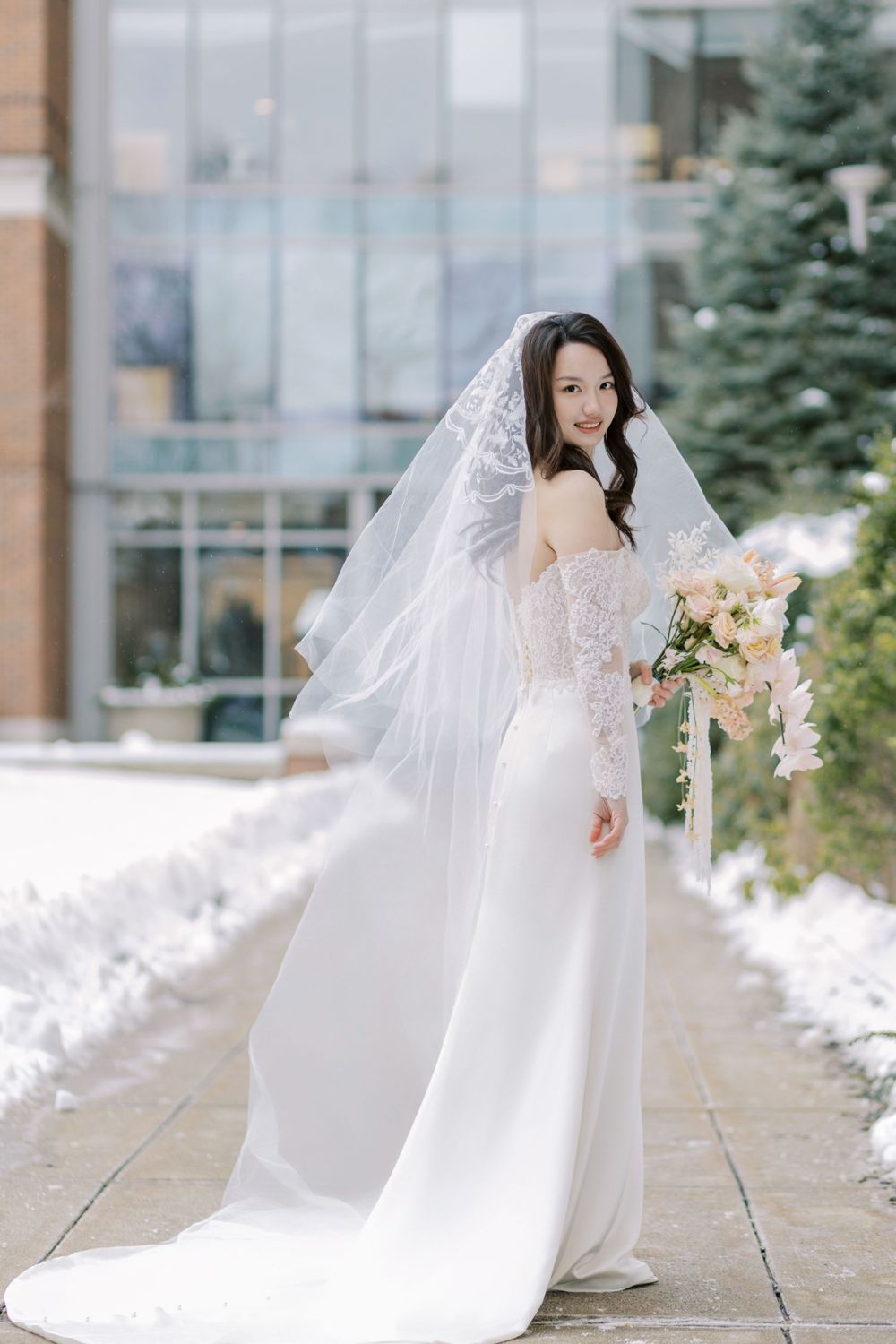 Bride in flowing lace gown poses on snowy pathway with bouquet.