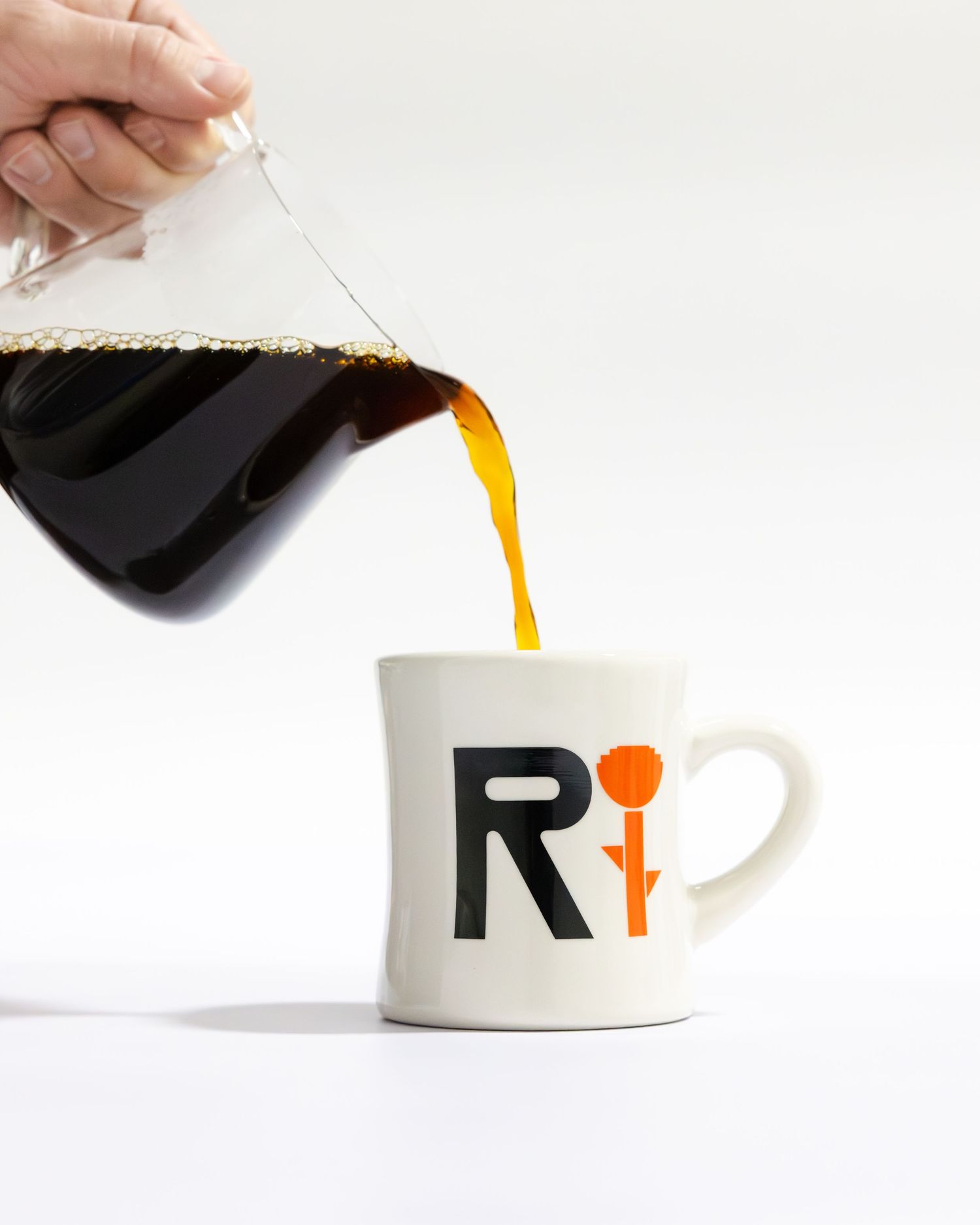 Coffee pouring from a glass carafe into a white ceramic mug with a black R logo design.