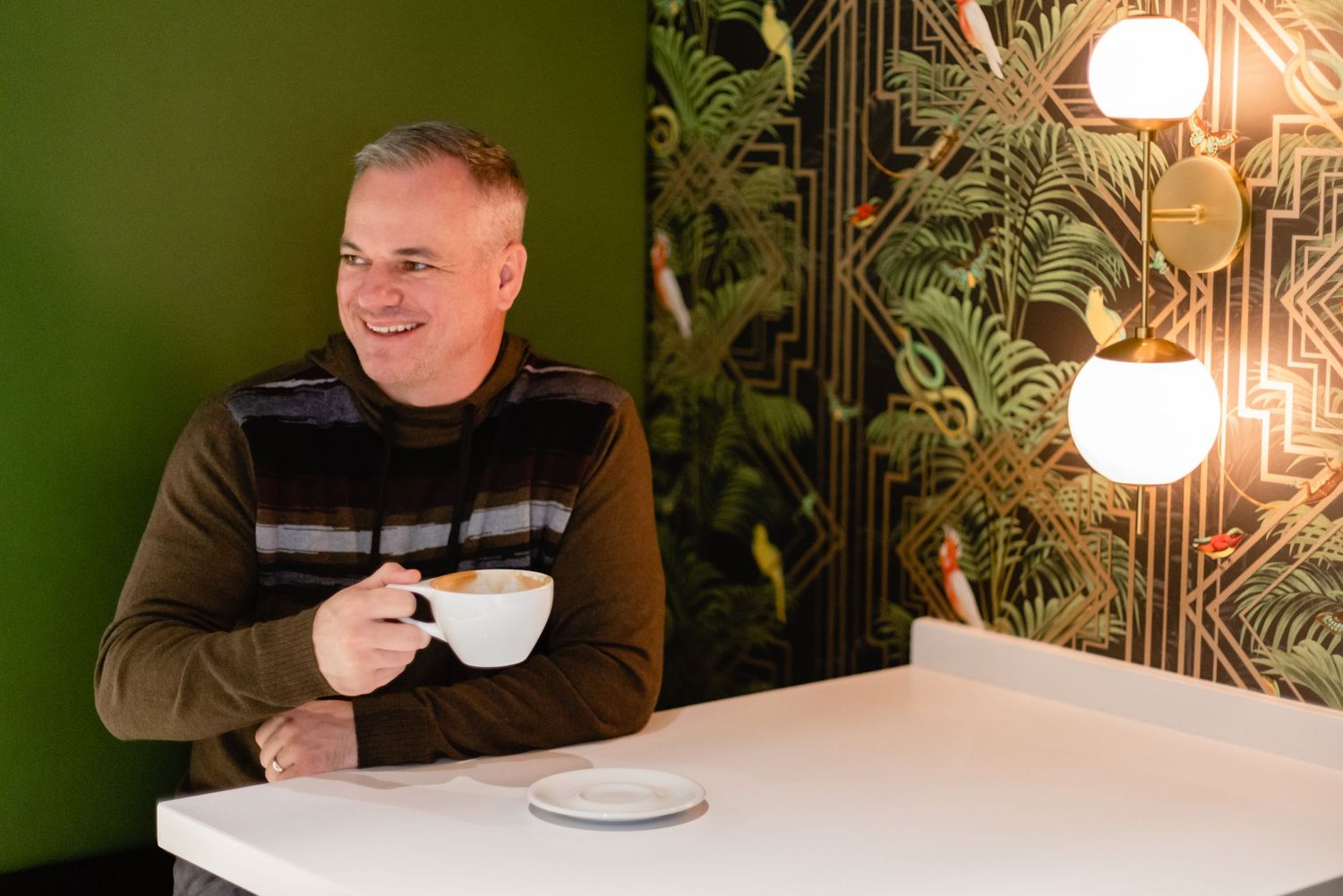 A person enjoys a warm beverage while sitting at a white table against a green wall with tropical wallpaper and lighting.