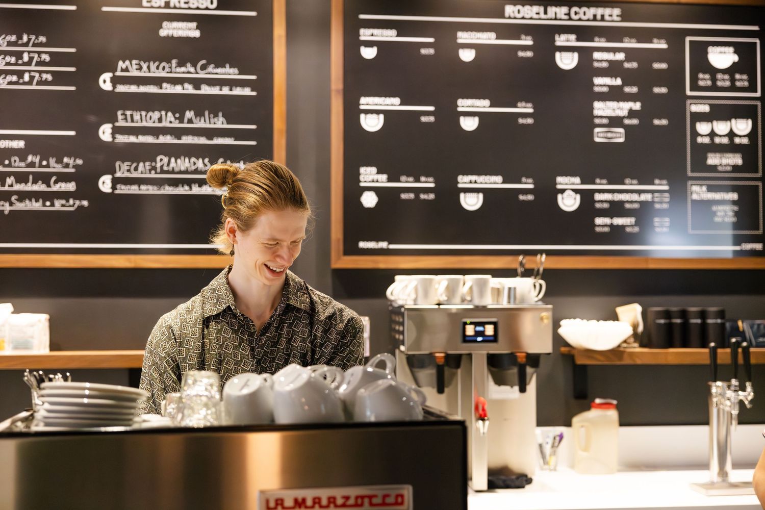 A barista works behind the counter at a coffee shop with a menu board displaying drink options in the background.