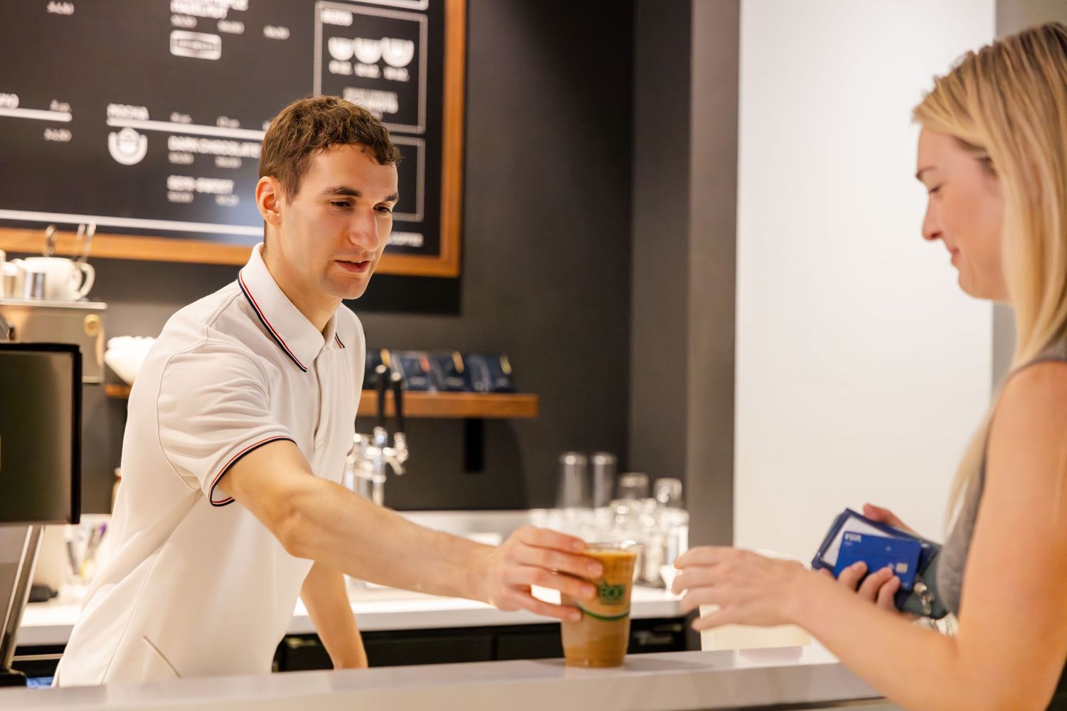 A bartender serves a customer at a bar counter with a dark wall and bottles visible behind.