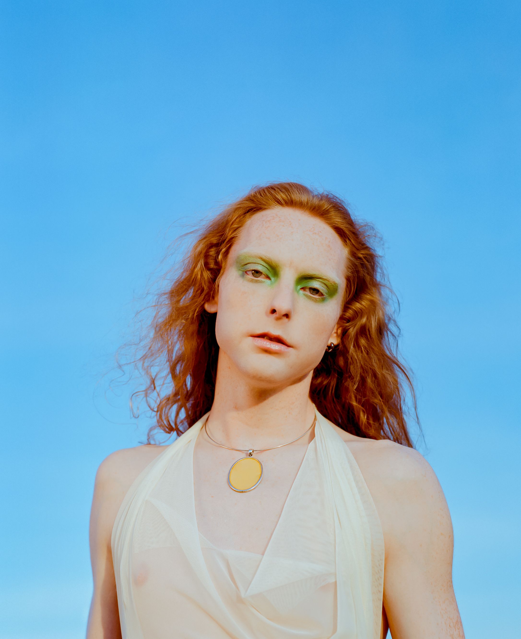 Portrait against blue sky featuring dramatic makeup and golden pendant.