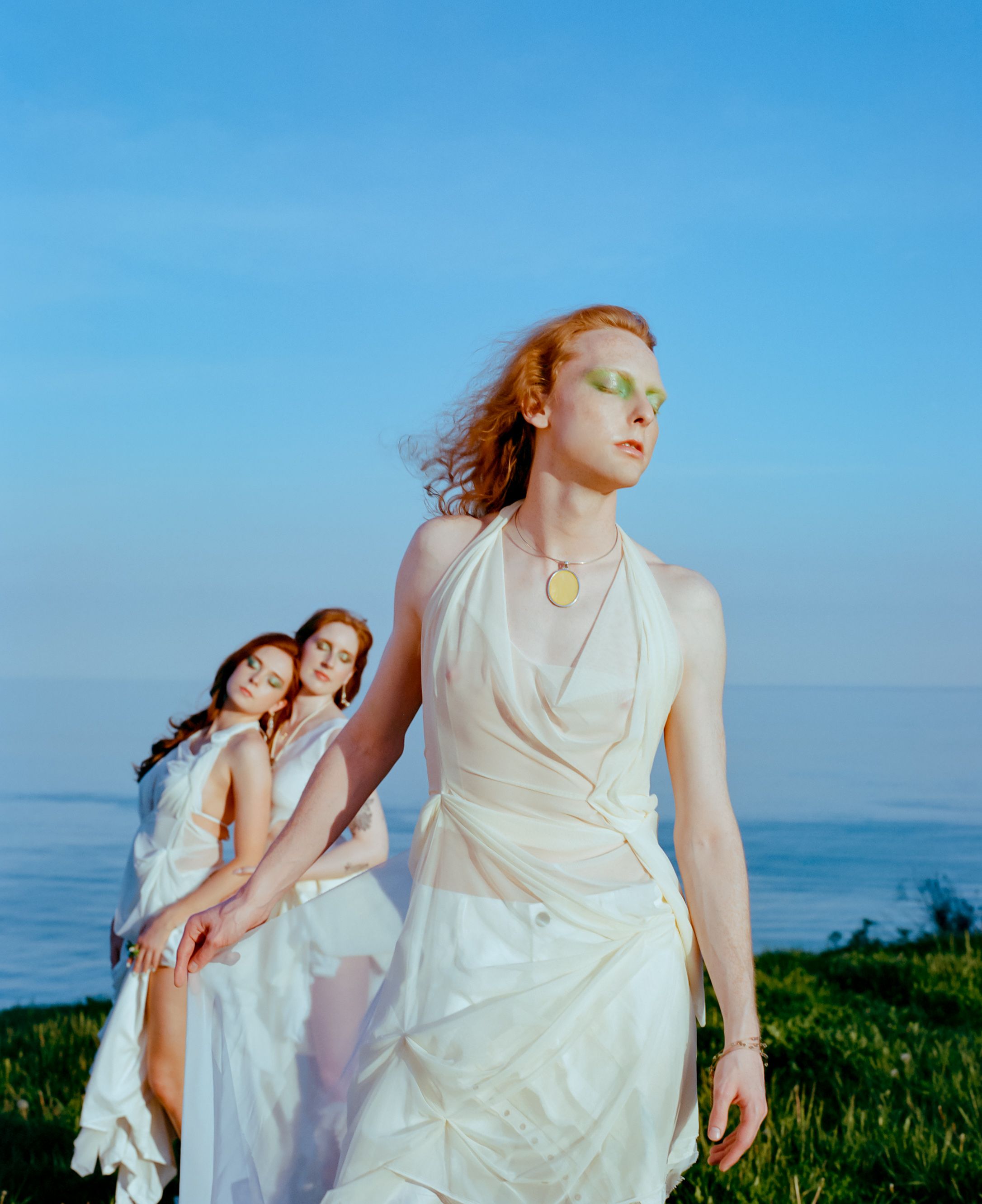 Three figures in flowing white dresses pose dramatically against a seaside landscape at dusk.