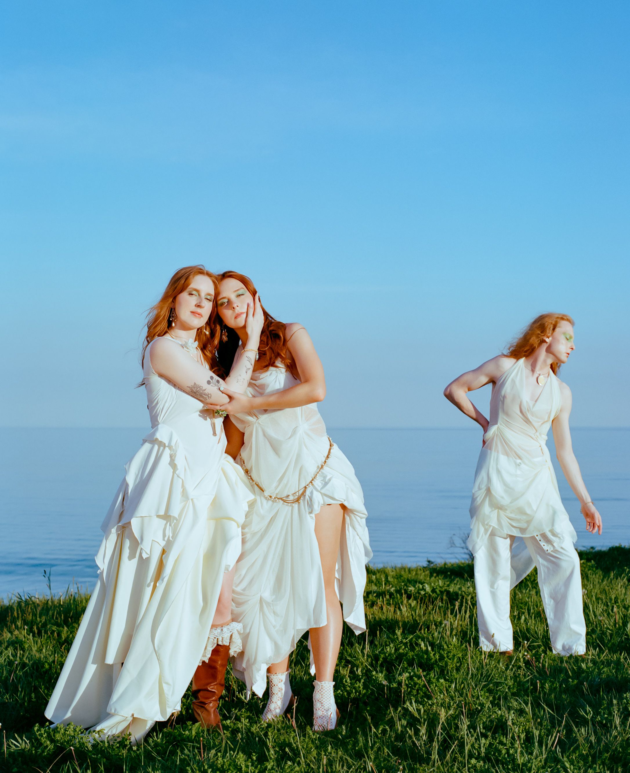 Three people in white outfits embrace and dance on a grassy cliff overlooking the ocean.