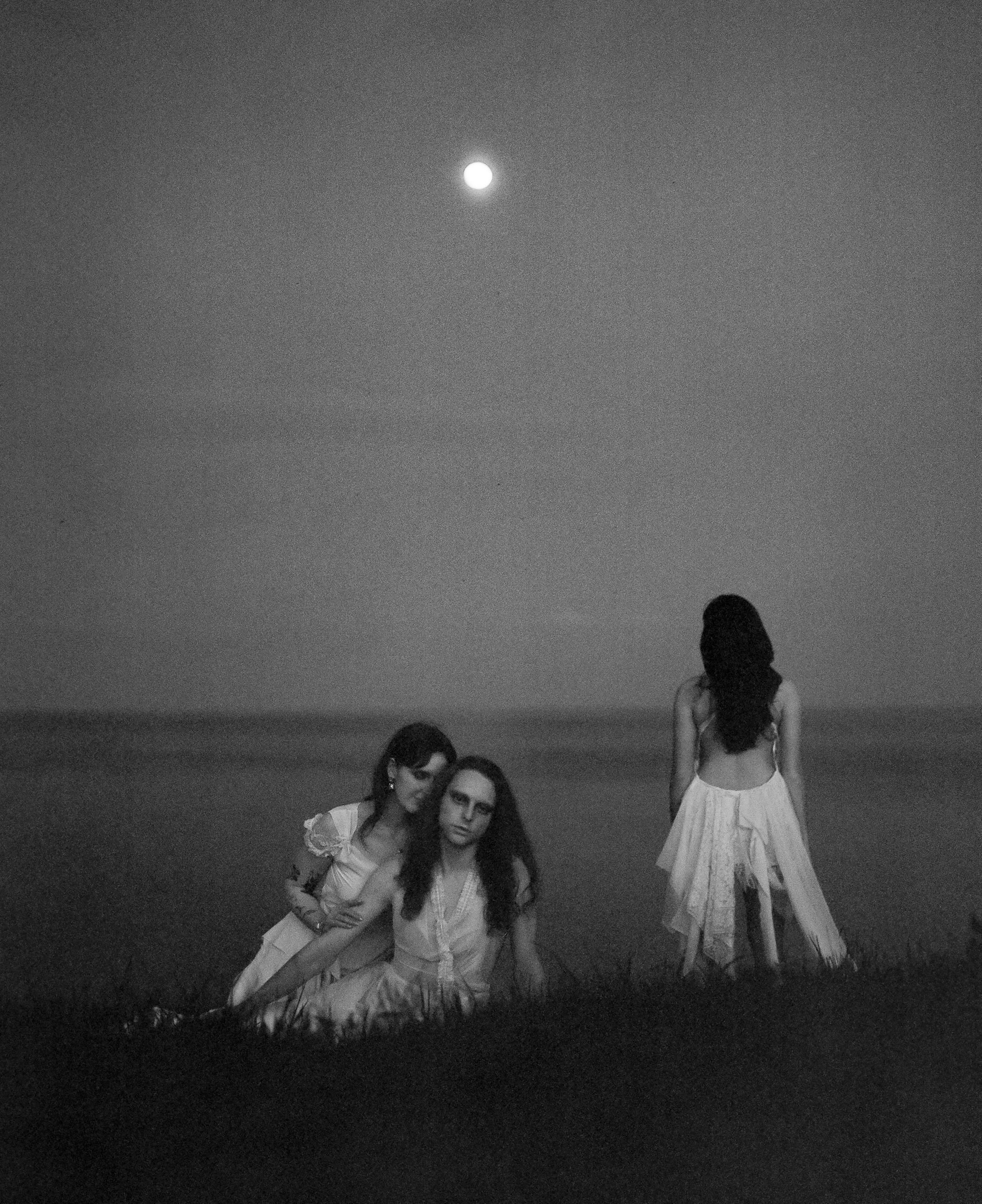Three silhouettes in white dresses stand under moonlight on a coastal hillside.