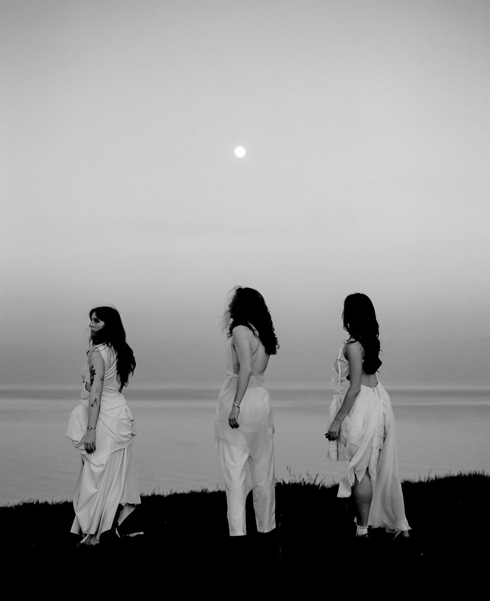 Black and white photo of three figures in white dresses standing in silhouette against a misty horizon.
