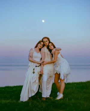 Three women embrace in ethereal white dresses against a dusky ocean backdrop.