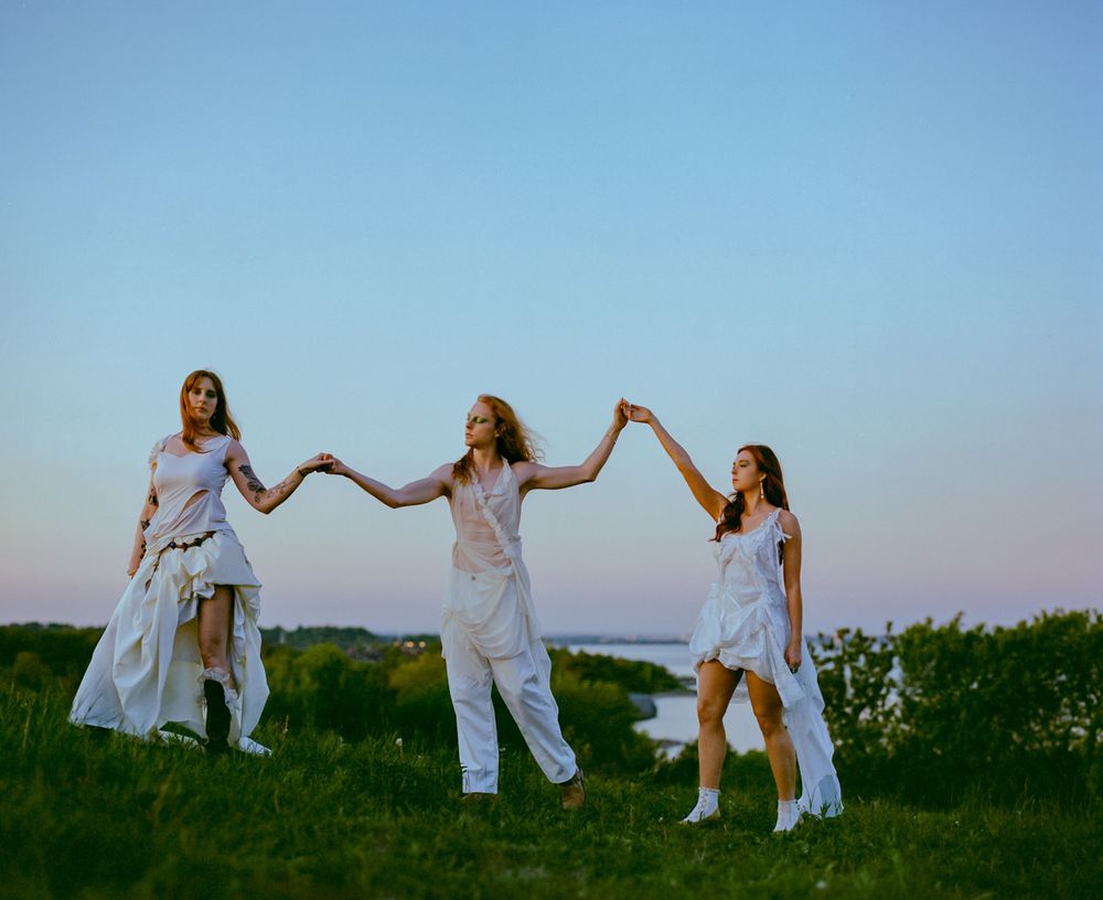 Three people in flowing white outfits hold hands while dancing on a hillside at sunset.
