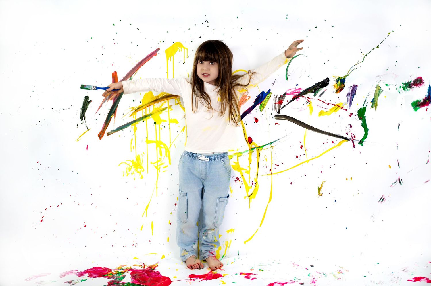 Someone stands against a white background with colorful paint splatters while wearing jeans and a light sweater.