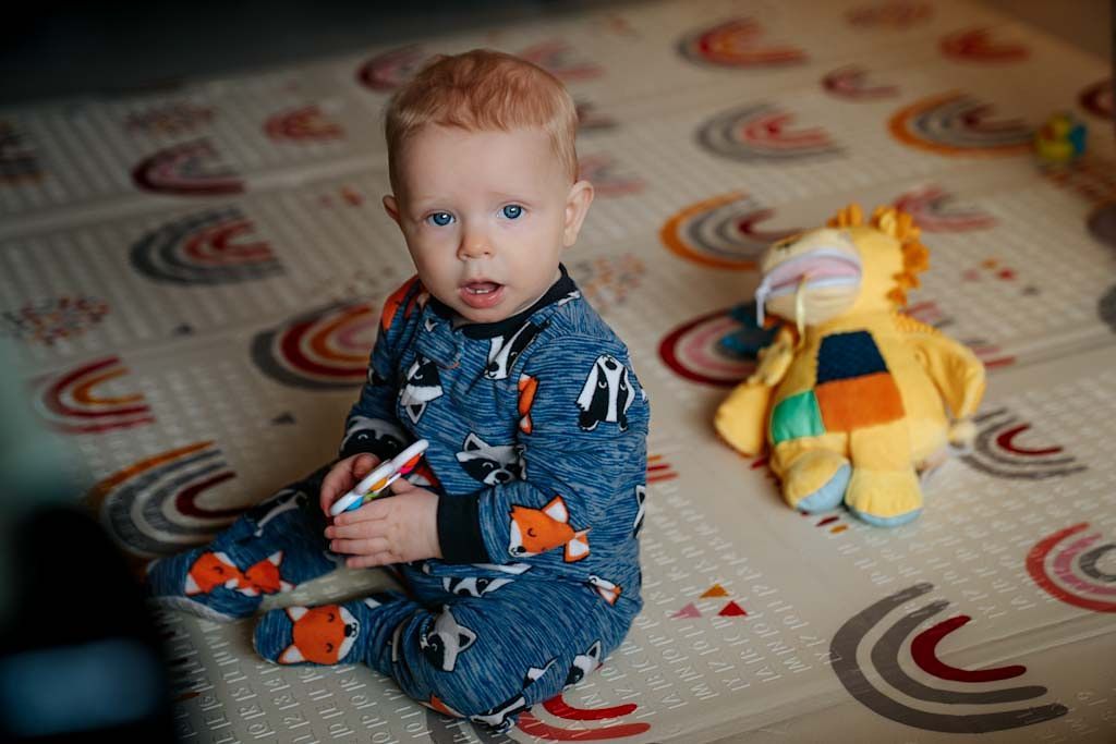 Adorable red-haired baby in dinosaur pajamas sitting on colorful play mat next to stuffed giraffe toy.