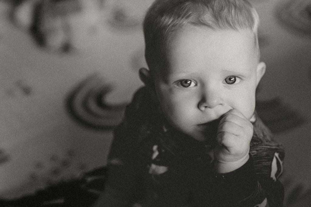 Black and white portrait of a cute baby with finger in mouth, gazing at camera on patterned surface.