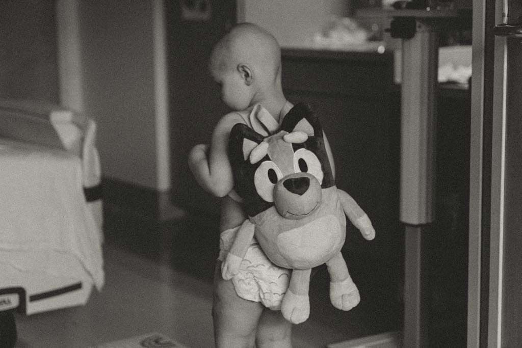 Black and white photo of a toddler in diaper carrying a Bluey stuffed toy walking through a doorway.