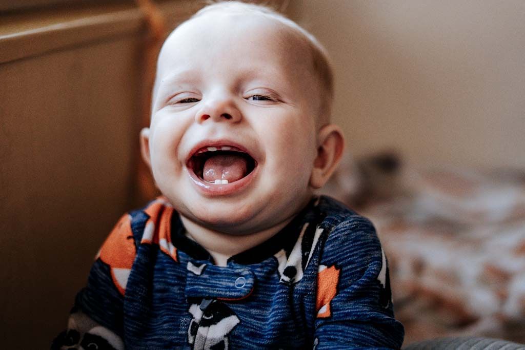 Happy laughing baby in dinosaur onesie showing first teeth, sitting in a warm cozy indoor setting.