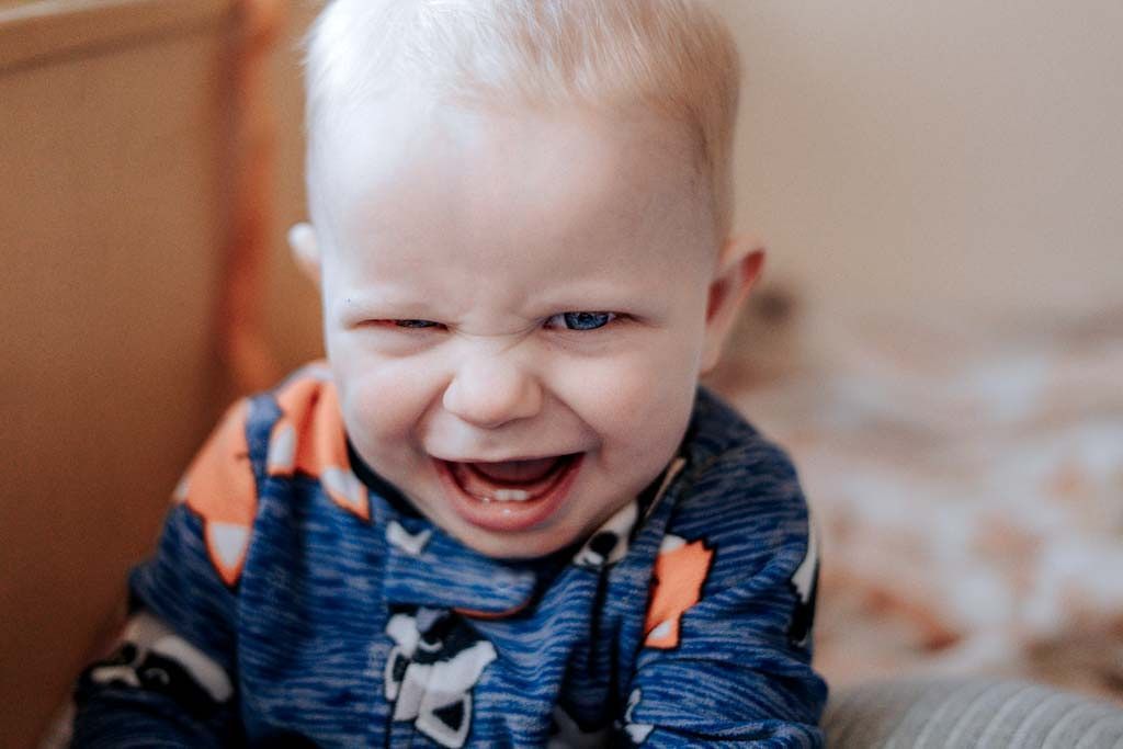 Joyful baby scrunching face in a big smile wearing a colorful dinosaur onesie in a cozy indoor setting.