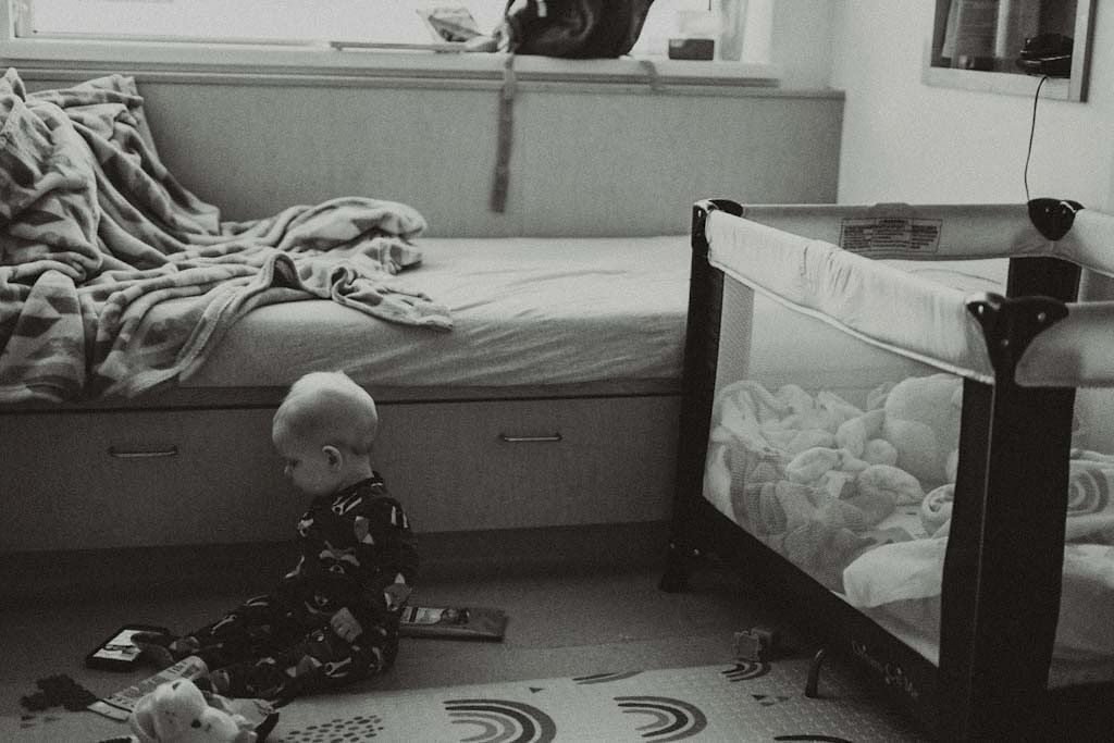 Black and white photo of a baby sitting on floor near a playpen.