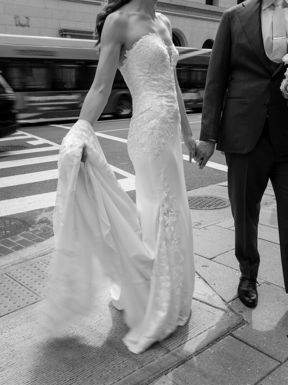 A black and white photo showcasing the intricate details of a fitted lace wedding gown on a city street.