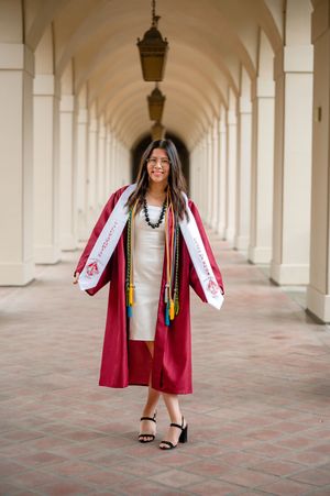 A graduate in academic regalia poses in an arched colonnade wearing a red graduation gown and white dress.