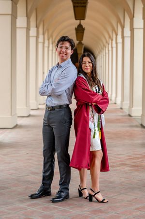 Graduates pose in formal attire under arched colonnade walkway with white columns and brick floor.