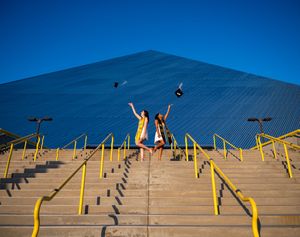 Two people jumping for joy on concrete steps with yellow handrails against a large blue pyramid structure.